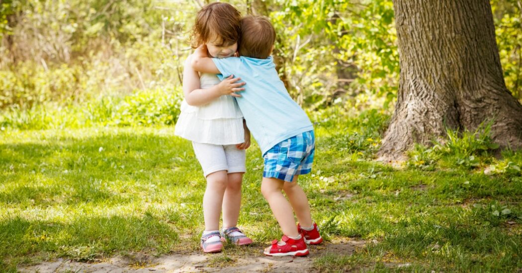 Two young children hug each other outside amongst some trees.